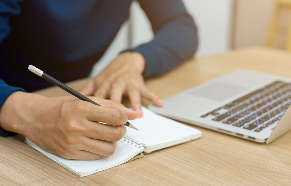 Person writing on a notebook beside a laptop